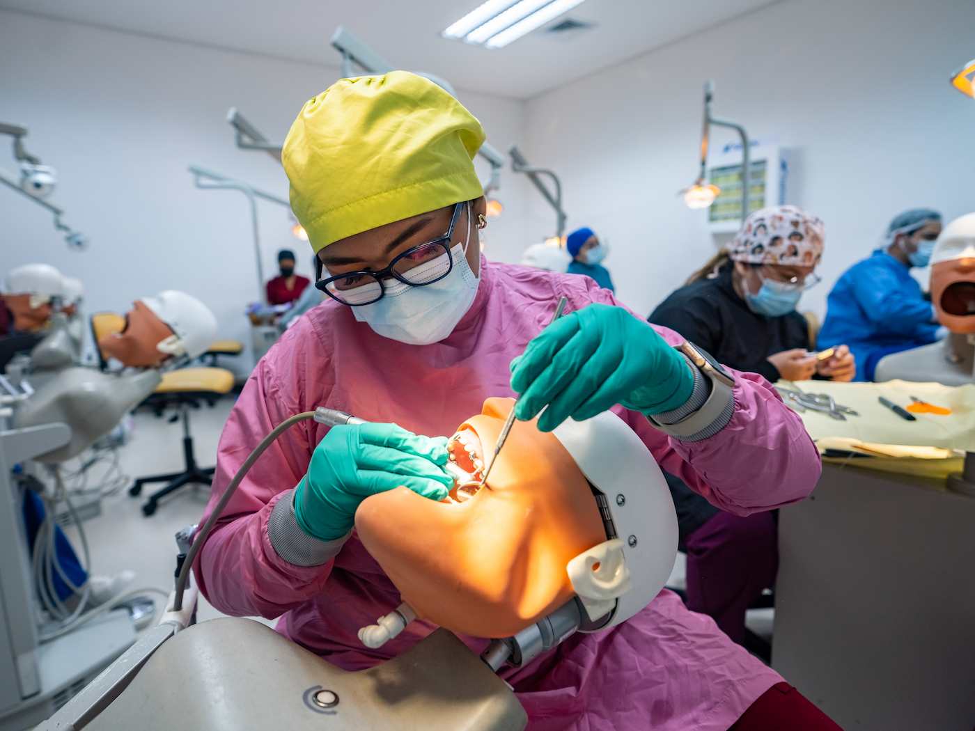 Estudiante de odontología realiza un procedimiento en un simulador de cabeza con boca abierta, usando instrumentos dentales en un aula práctica.